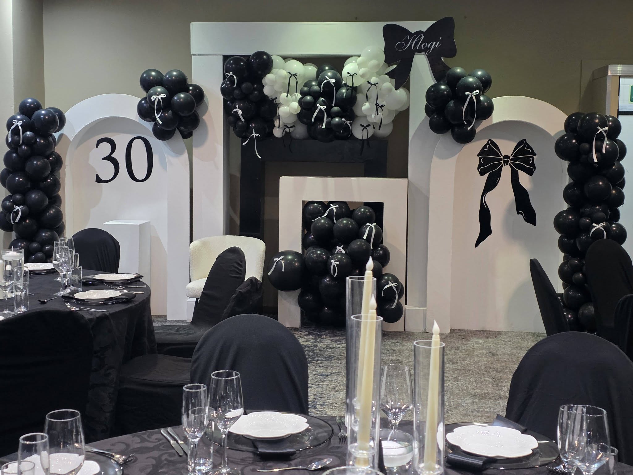 30th birthday party setup at Muano, black and white balloon arch, fireplace prop and round tables with dark tablecloths